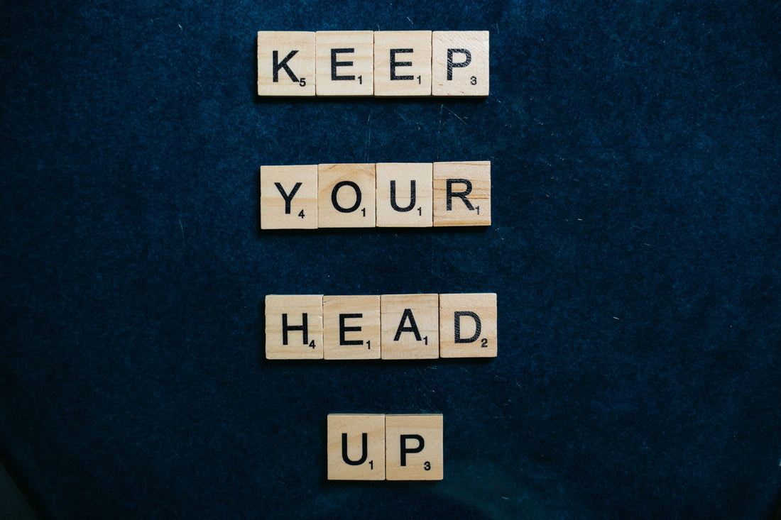 Wooden Scrabble letter tiles spelling “KEEP YOUR HEAD UP” arranged in four rows against a dark navy-blue textured background.